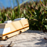 Close up of the Candylab wooden diecast campervan toy on some light sand in front of some green leaves
