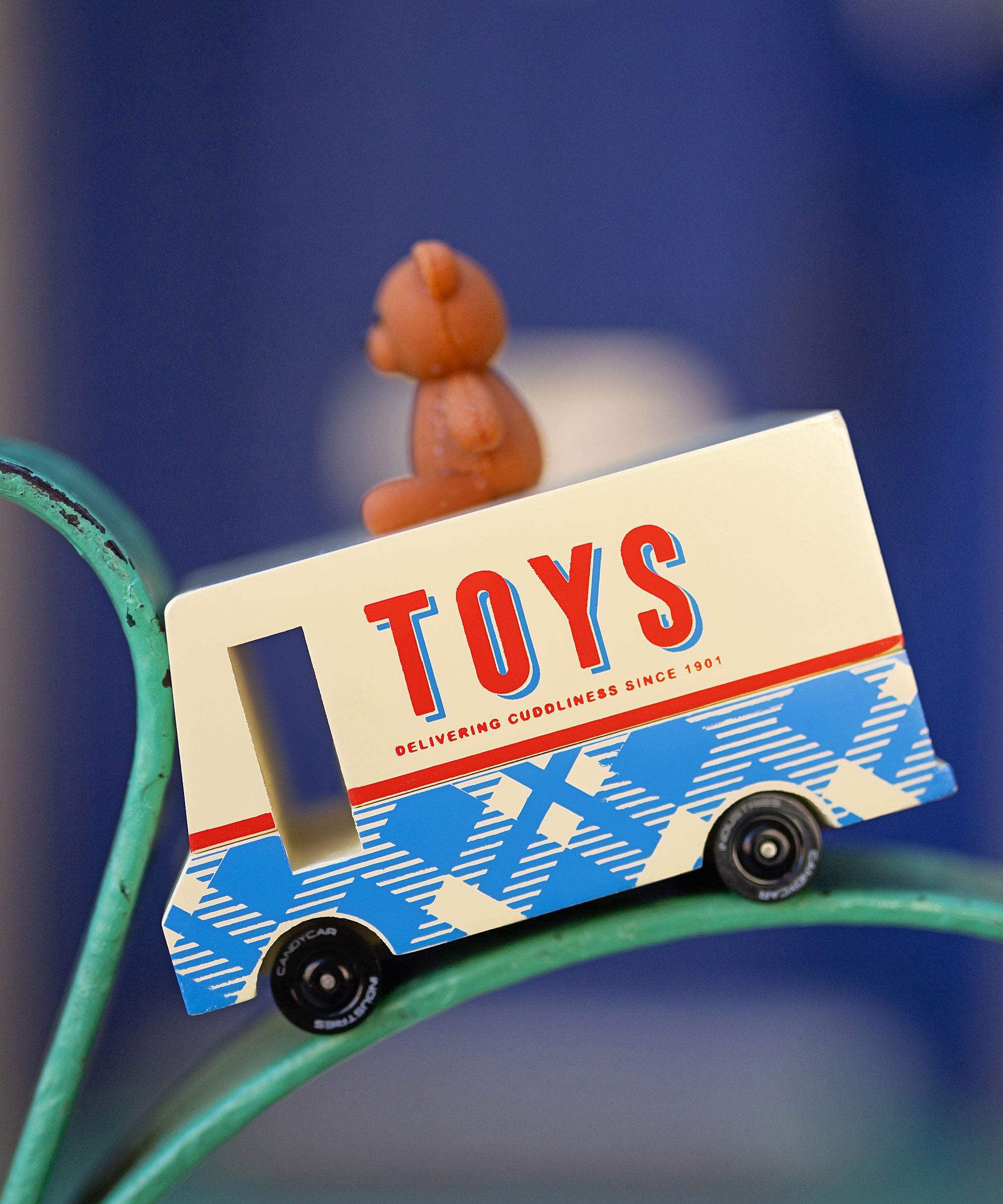 CandyLab cream and blue CandyVan with a brown teddy bear on the roof with Toys text logo in red on the side - placed on a chair frame with a faded blue background.