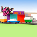 Candylab handmade wooden racing car and pink sedan toys towing wooden caravans into a toy motel model on a green and blue background