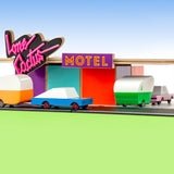 Candylab handmade wooden racing car and pink sedan toys towing wooden caravans into a toy motel model on a green and blue background