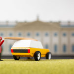 Toy figure putting a miniature surfboard on top of the candylab gold cotswold toy vehicle in front of a mansion model