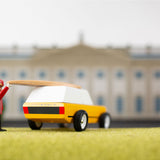 Toy figure putting a miniature surfboard on top of the candylab gold cotswold toy vehicle in front of a mansion model
