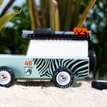 Side of the candylab wooden zebra drifter vehicle toy on some sand in front of a green plant