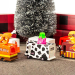 6 wooden Candylab toy trucks lined up on a brown carpet in front of a Christmas tree and some red presents