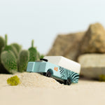 Close up of the Candylab wooden SUV toy on some sand in front of a stack of rocks and a miniature cactus