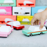 Close up of a childs hand holding the Candylab handmade teal wagon toy vehicle next to the pink sedan and taxi toys