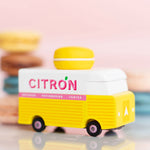 Picture of the Citron Candylab Candyvan. It is a 1960s style yellow and white van with a yellow Lemon Macaron on the roof. The background shows colourful macarons.