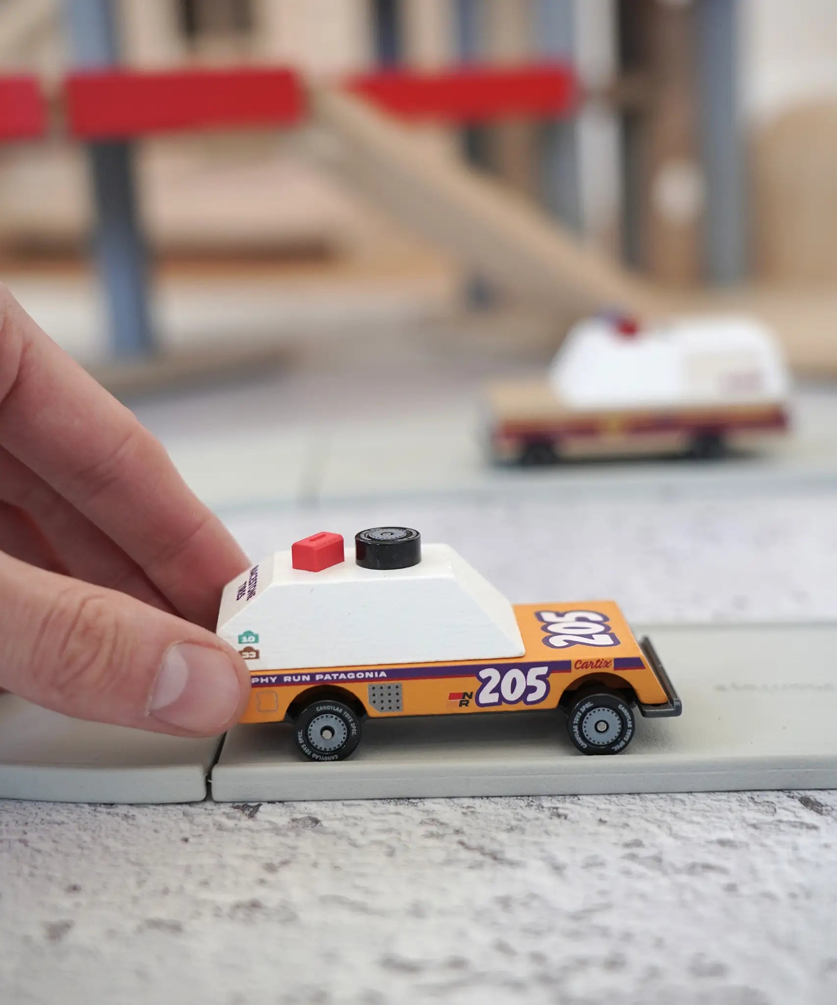 Adult hand pushing a orange Candylab rust patrol car on a play road showing scale 