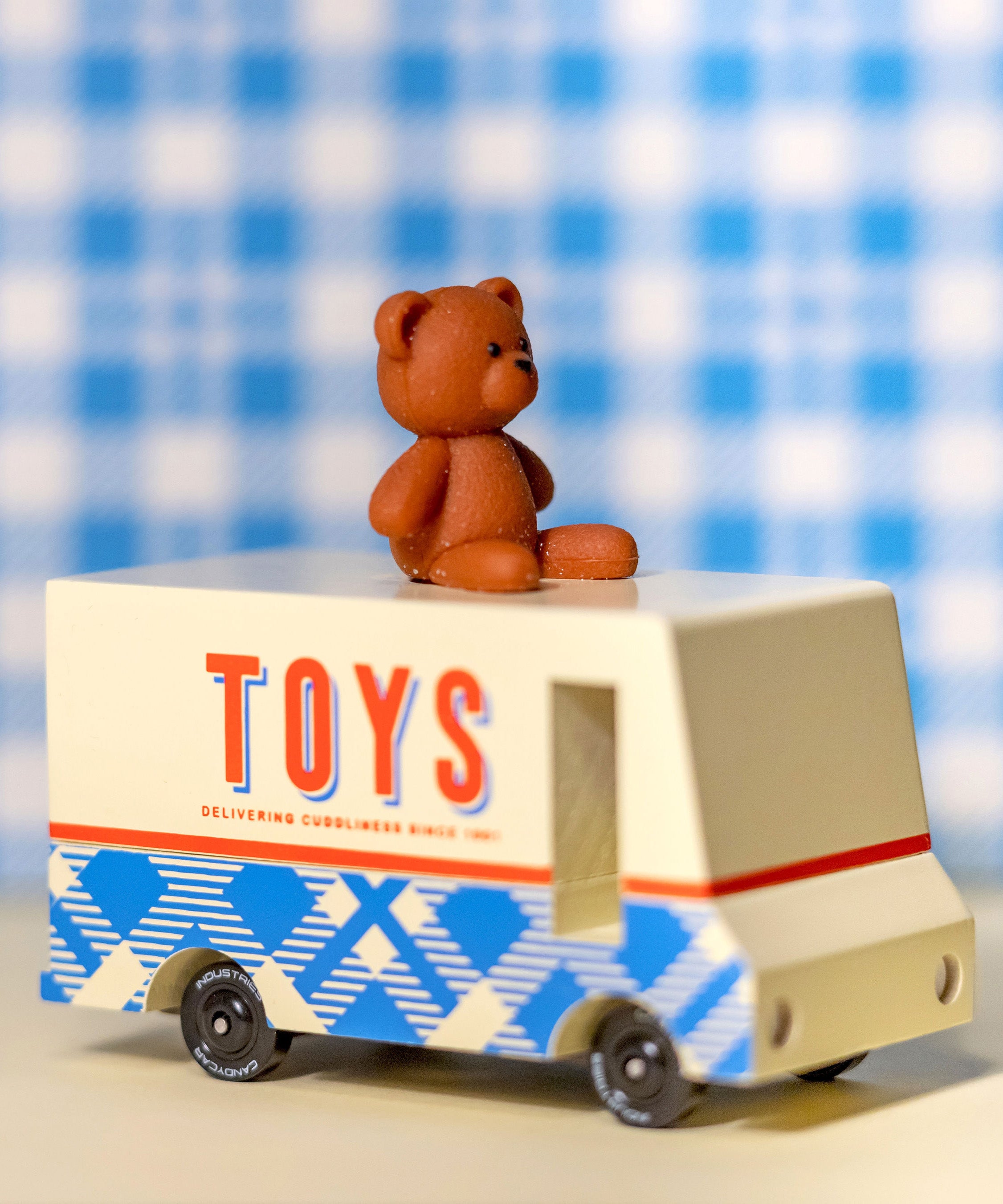 CandyLab cream and blue CandyVan with a brown teddy bear on the roof with Toys text logo in red on the side - on a table with a blue check blanket in the background.