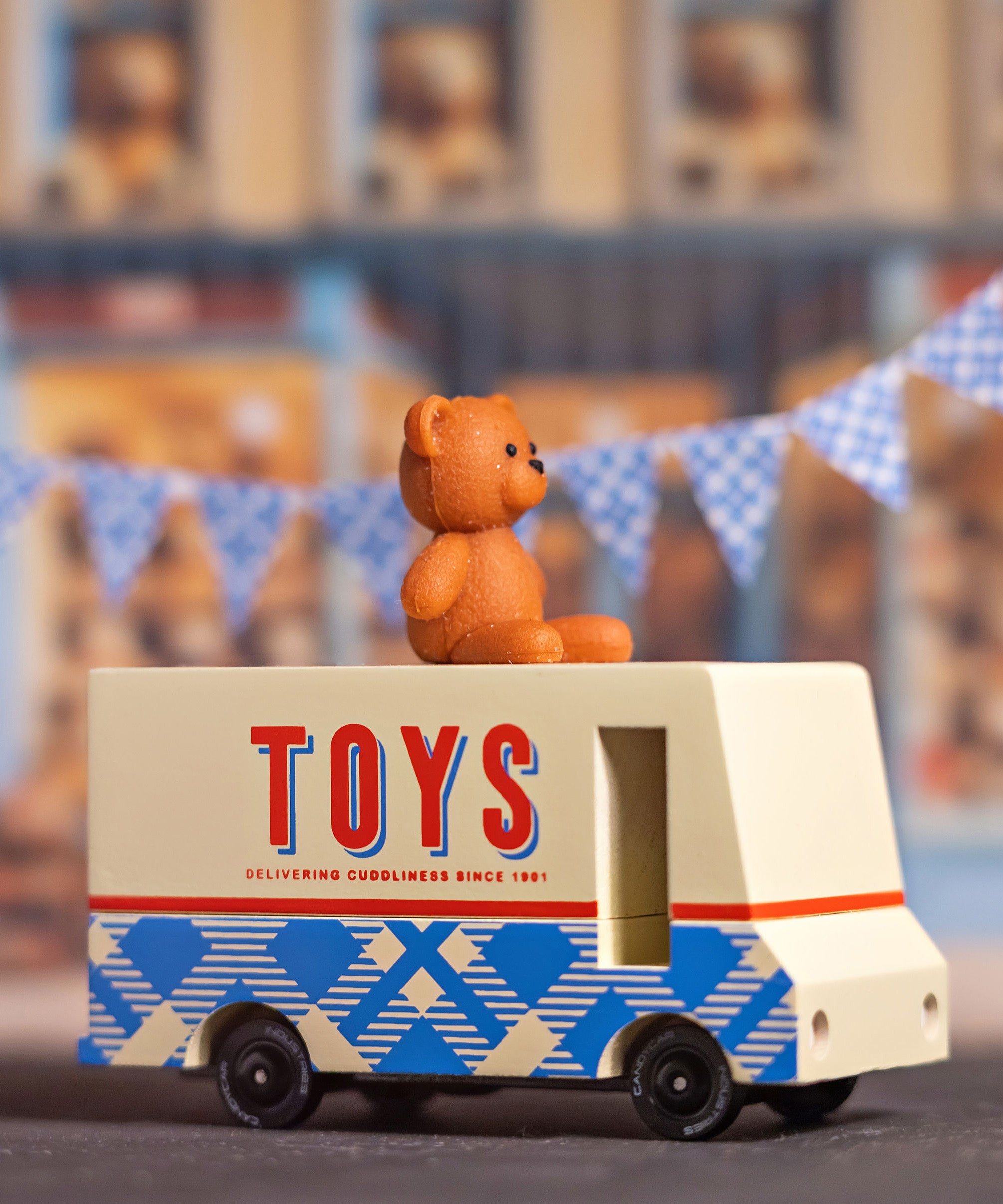 CandyLab cream and blue CandyVan with a brown teddy bear on the roof with Toys text logo in red on the side - with blue check bunting in the background.
