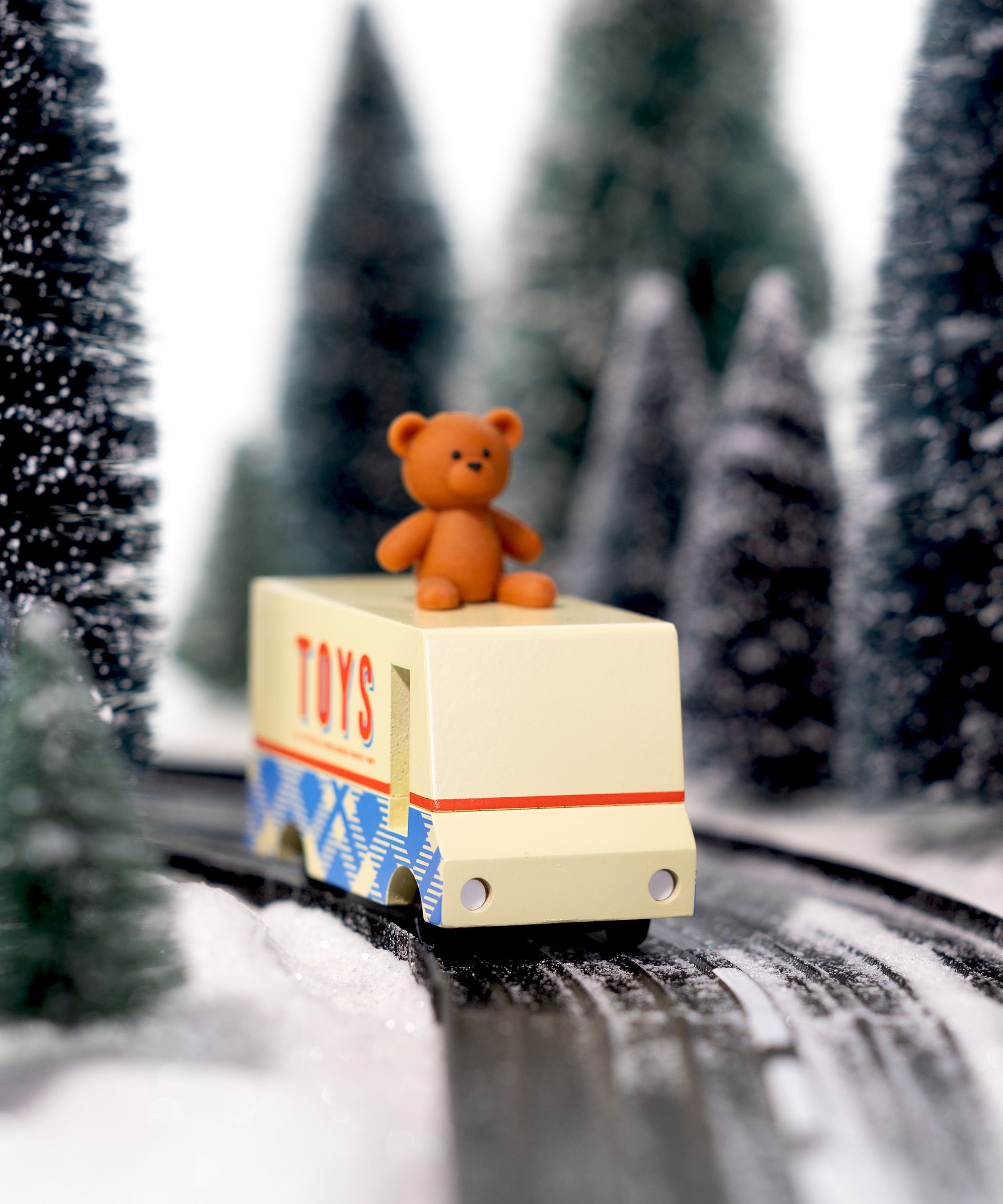 CandyLab cream and blue CandyVan with a brown teddy bear on the roof with Toys text logo in red on the side - on a road going through a snowy forest.