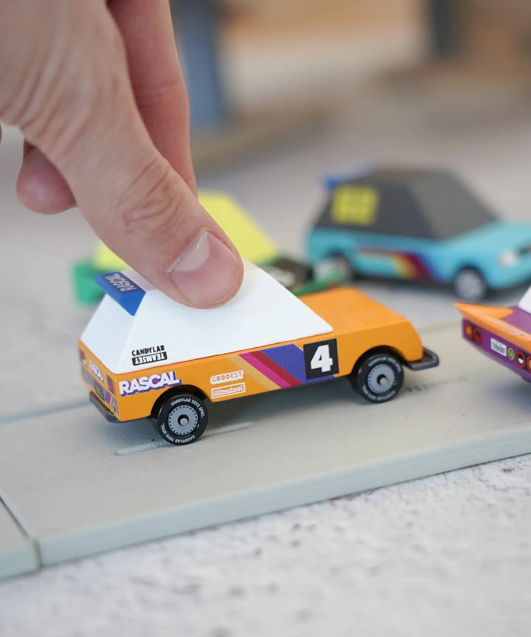 Adult hand holding a orange racing car from the Candylab Tokyo heat toon cars set on a play road showing scale