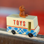 CandyLab cream and blue CandyVan with a brown teddy bear on the roof with Toys text logo in red on the side - on a chair frame with a blurred red background.