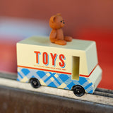 CandyLab cream and blue CandyVan with a brown teddy bear on the roof with Toys text logo in red on the side - on a chair frame with a blurred red background.