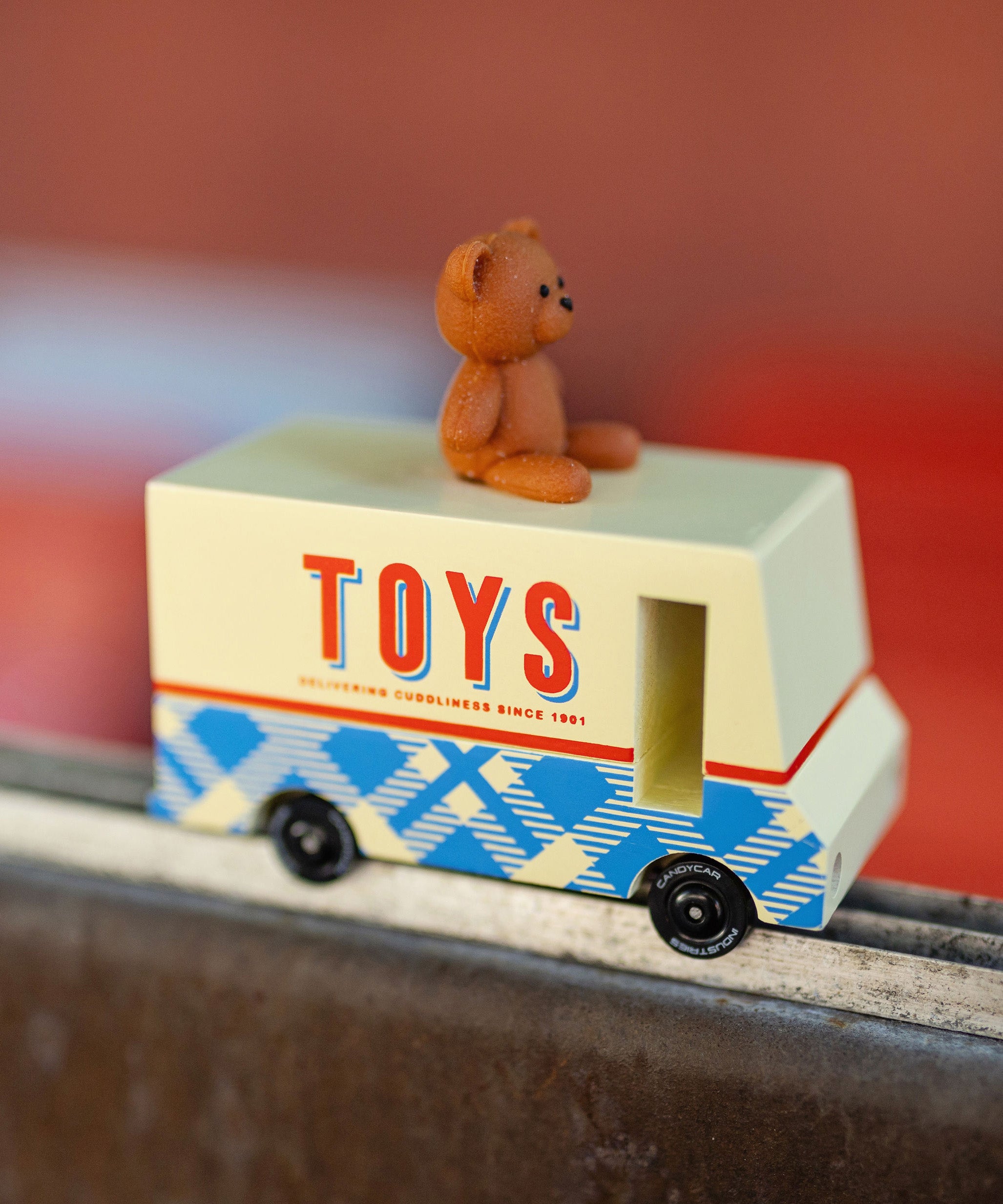 CandyLab cream and blue CandyVan with a brown teddy bear on the roof with Toys text logo in red on the side - on a chair frame with a blurred red background.