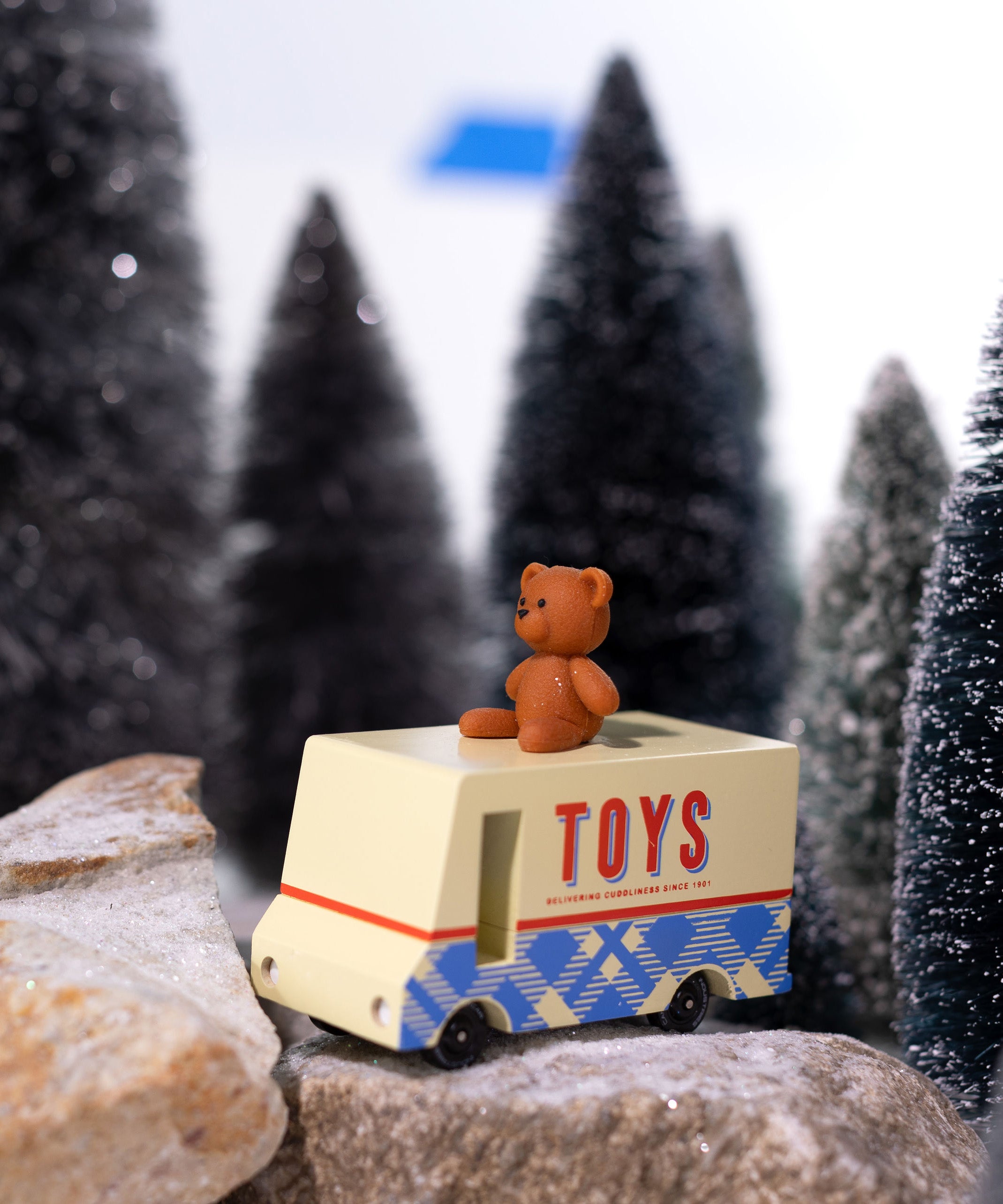 CandyLab cream and blue CandyVan with a brown teddy bear on the roof with Toys text logo in red on the side - parked on rocks with a snowy forest in the background.