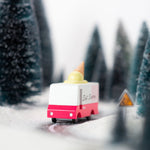 Close up of the Candylab wooden ice cream van toy on a black toy road surrounded by small trees and snow