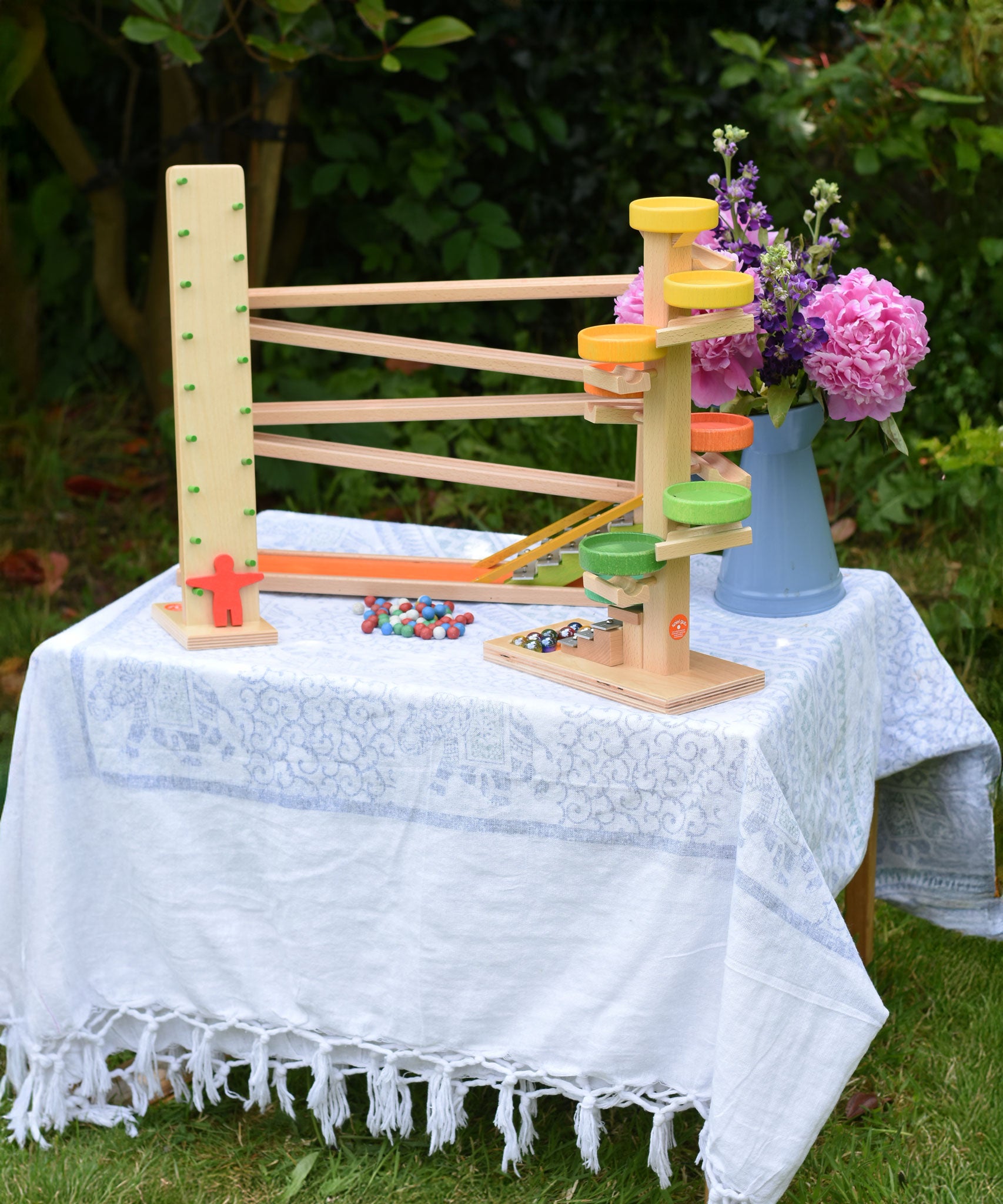 A collection of Beck Spielwaren wooden toys placed on a small table outdoors. The Wobbler tumbling toy, the run plate chimes marble run tower and the chimes marble run track. 