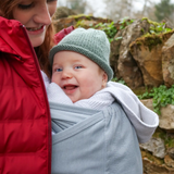 Close Caboo Lite Baby Carrier in Alloy shown outdoors with a smiling baby carried close against the wearer’s chest.