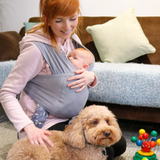 Close Caboo Lite Baby Carrier in Alloy with a newborn baby in ergonomic front carry position while the mother sits with a dog in a living room. 