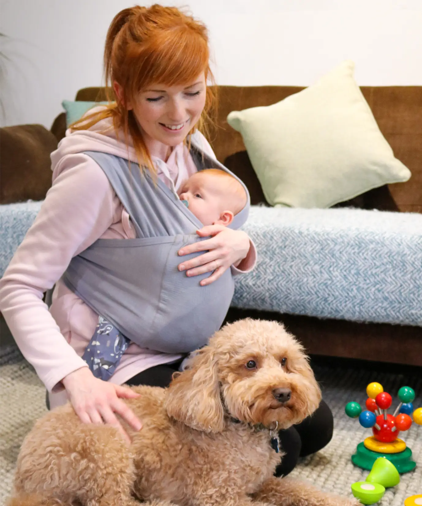 Close Caboo Lite Baby Carrier in Alloy with a newborn baby in ergonomic front carry position while the mother sits with a dog in a living room. 