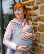 Woman leaning against a stone wall holding a baby in the close caboo lite baby carrier in the light grey alloy colour