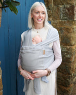 Woman stood in front of a blue door wearing the Close caboo organic baby carrier in the porpoise colour