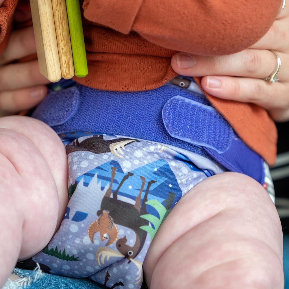 A baby wearing The Close Parent Pop-in reusable nappy wrap cover with velcro closures, in a purple moose print