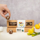 Cocoa Loco fairtrade hot cross bun easter chocolate truffles boxes and truffle being held in a persons fingers. On a white tables with colourful toys eggs