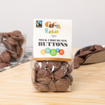 Bag of Cocoa Loco organic milk chocolate buttons on a wooden table in front of a glass cup and metal scoop filled with chocolate buttons