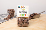 Bag of Cocoa Loco organic milk chocolate buttons on a wooden table in front of a glass cup and metal scoop filled with chocolate buttons