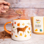 Close up of hand holding some Cocoa Loco organic hot chocolate flakes on a spoon above a white mug with a dog painting on the front