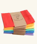 Six organic cotton play cloths by Laura's Play Cotton, in solid colours folded and stacked in rainbow order.