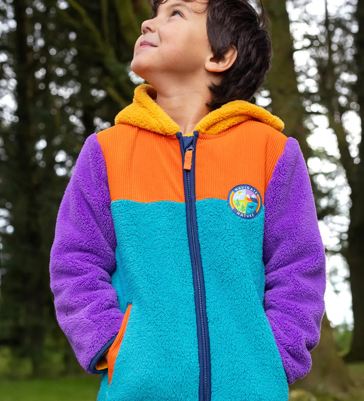 A child wearing a colorful fleece zip-up hoodie outdoors in a forest. The hoodie has a bright turquoise front, orange yoke, purple sleeves, and a yellow hood lining.