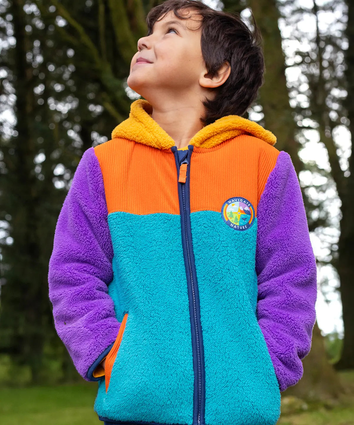 A child wearing a colorful fleece zip-up hoodie outdoors in a forest. The hoodie has a bright turquoise front, orange yoke, purple sleeves, and a yellow hood lining.