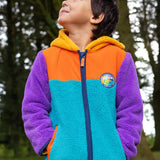 A child wearing a colorful fleece zip-up hoodie outdoors in a forest. The hoodie has a bright turquoise front, orange yoke, purple sleeves, and a yellow hood lining.