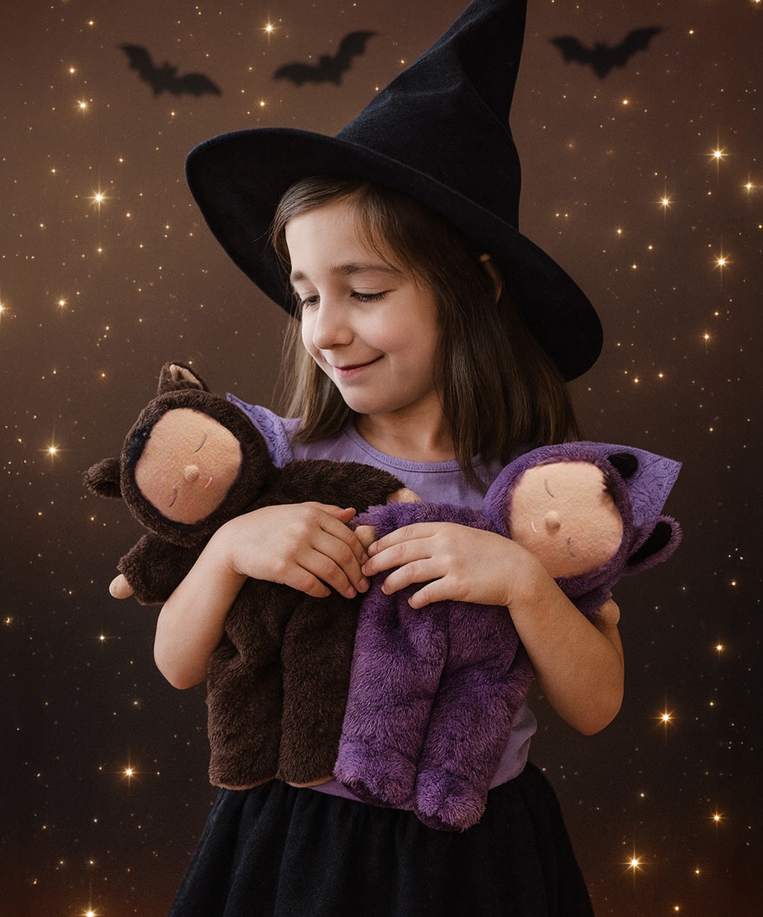 A child wearing a witches hat cuddling two Olli Ella cozy dozy bats in brown and purple. These soft toys are part of Olli Ella's Halloween collection available here at Babipur. 