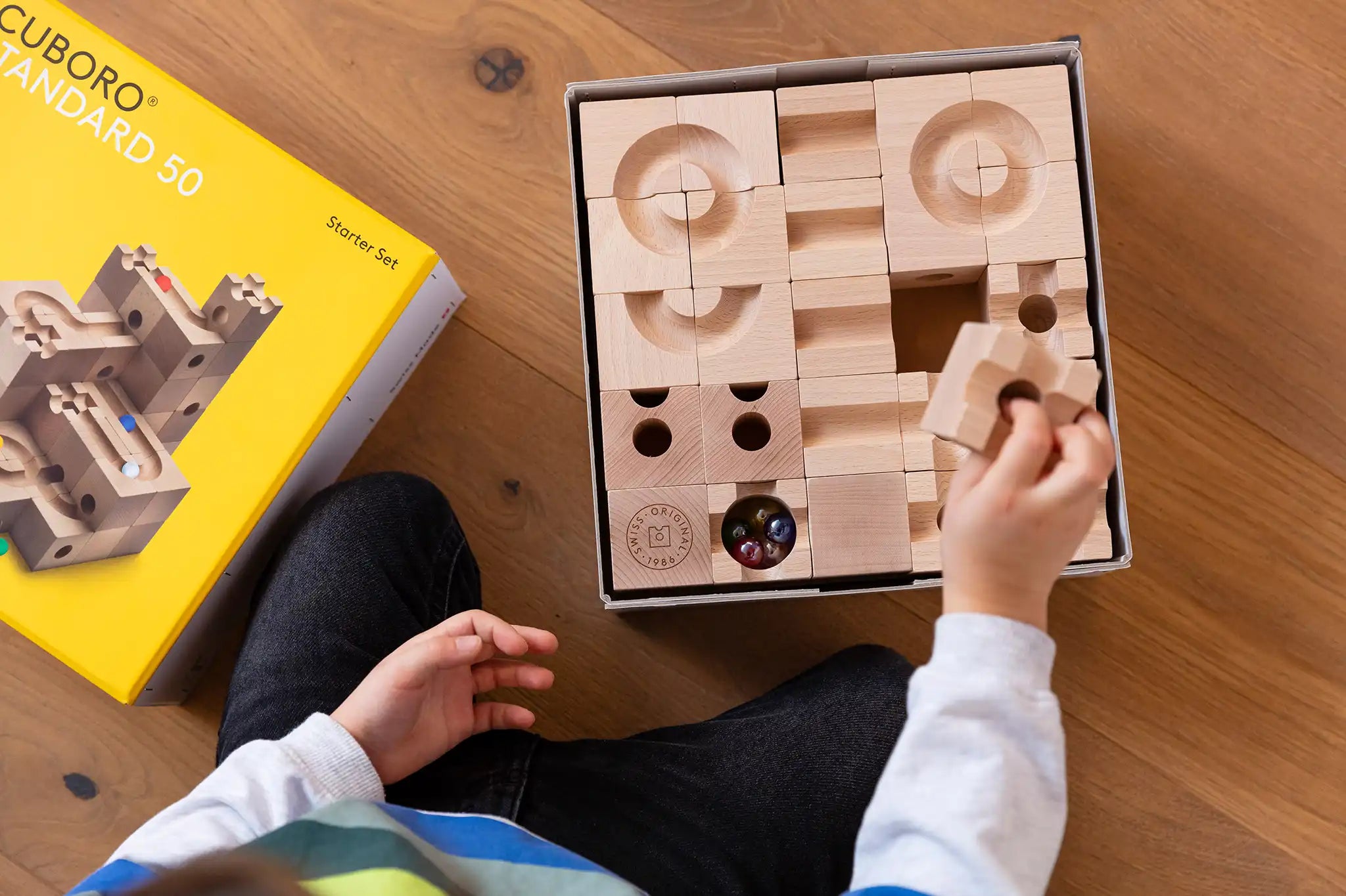 Cuboro standard 50 piece wooden marble run set in it's box, a child's hand can be seen taking a piece out of the box. 