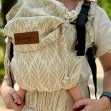 Front of mother wearing a Didymos didyfix full buckle baby carrier in Trias cream line showing repeating leaf design on cotton fabric