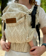 Front of mother wearing a Didymos didyfix full buckle baby carrier in Trias cream line showing repeating leaf design on cotton fabric