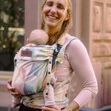 Mother carrying a baby in the zephyr  Didymos didyflow half buckle carrier in the frontal position. A cream carrier with repeating geometric design in a pastel palette on cotton fabric