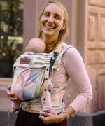 Mother carrying a baby in the zephyr  Didymos didyflow half buckle carrier in the frontal position. A cream carrier with repeating geometric design in a pastel palette on cotton fabric