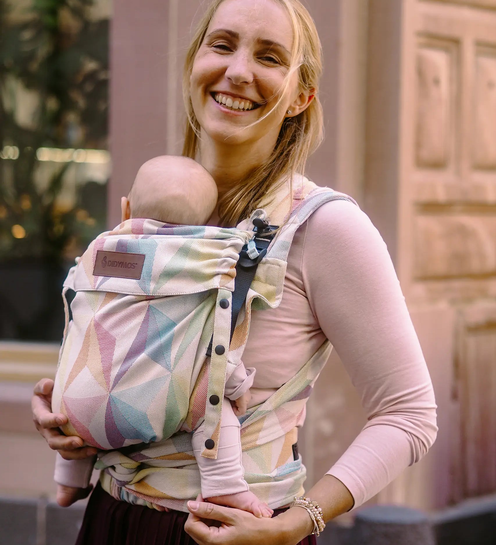Mother carrying a baby in the zephyr  Didymos didyflow half buckle carrier in the frontal position. A cream carrier with repeating geometric design in a pastel palette on cotton fabric