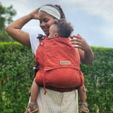 Mother wearing a Didymos didyfix full buckle toddler carrier in rusty red showing a toddler sitting inside