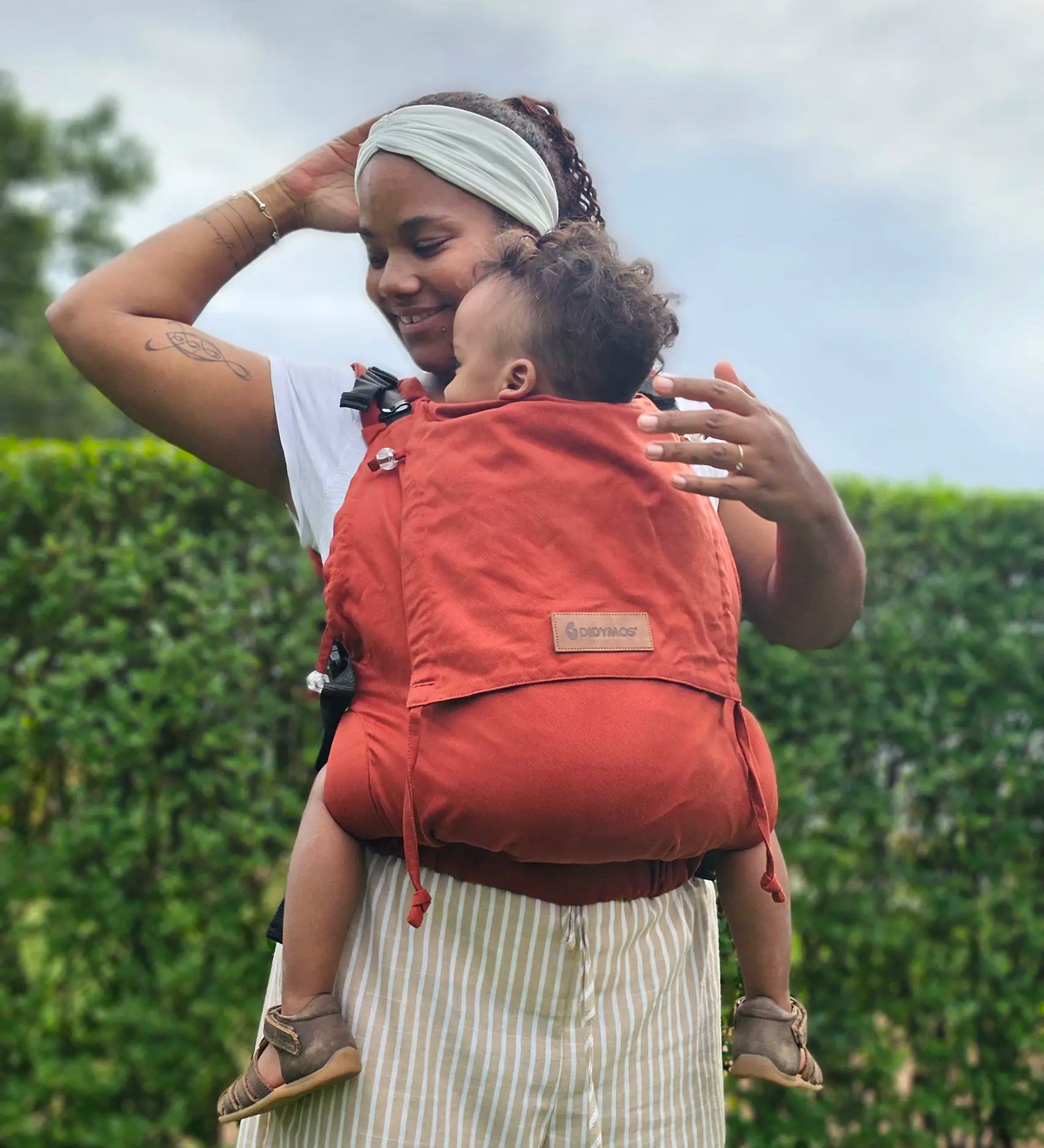 Mother wearing a Didymos didyfix full buckle toddler carrier in rusty red showing a toddler sitting inside