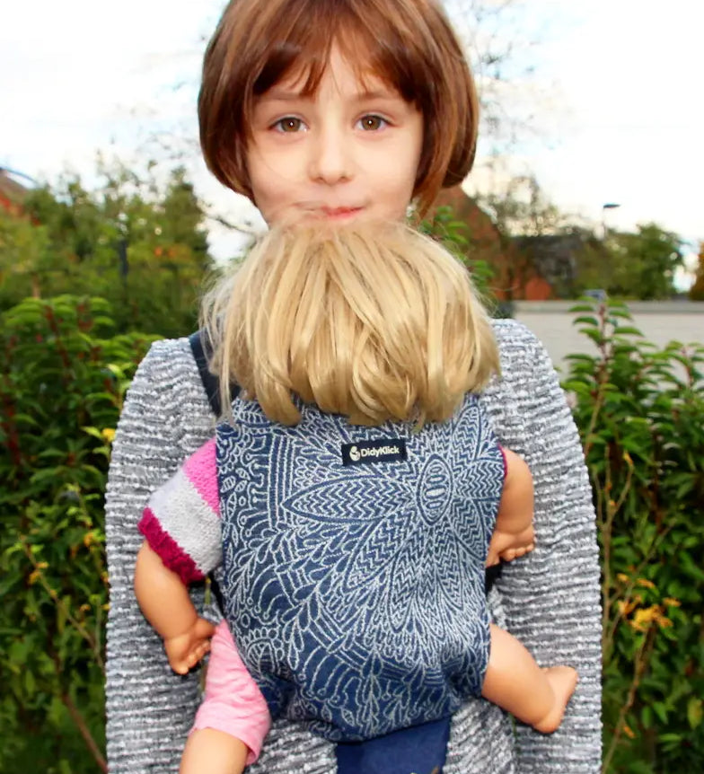Child carrying a doll in a Didymos toy cotton carrier in the blue and white Kipos design in the frontal position