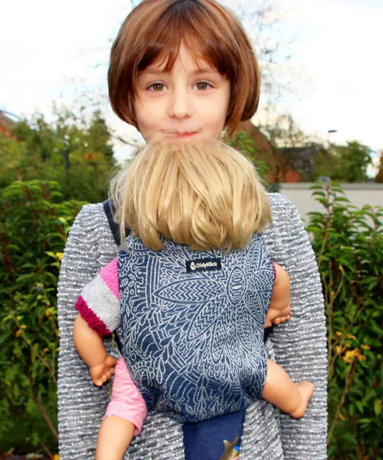 Child carrying a doll in a Didymos toy cotton carrier in the blue and white Kipos design in the frontal position