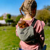Child carrying a doll in a Didymos toy cotton carrier in olive twig. A green carrier with a repeating tree design. 