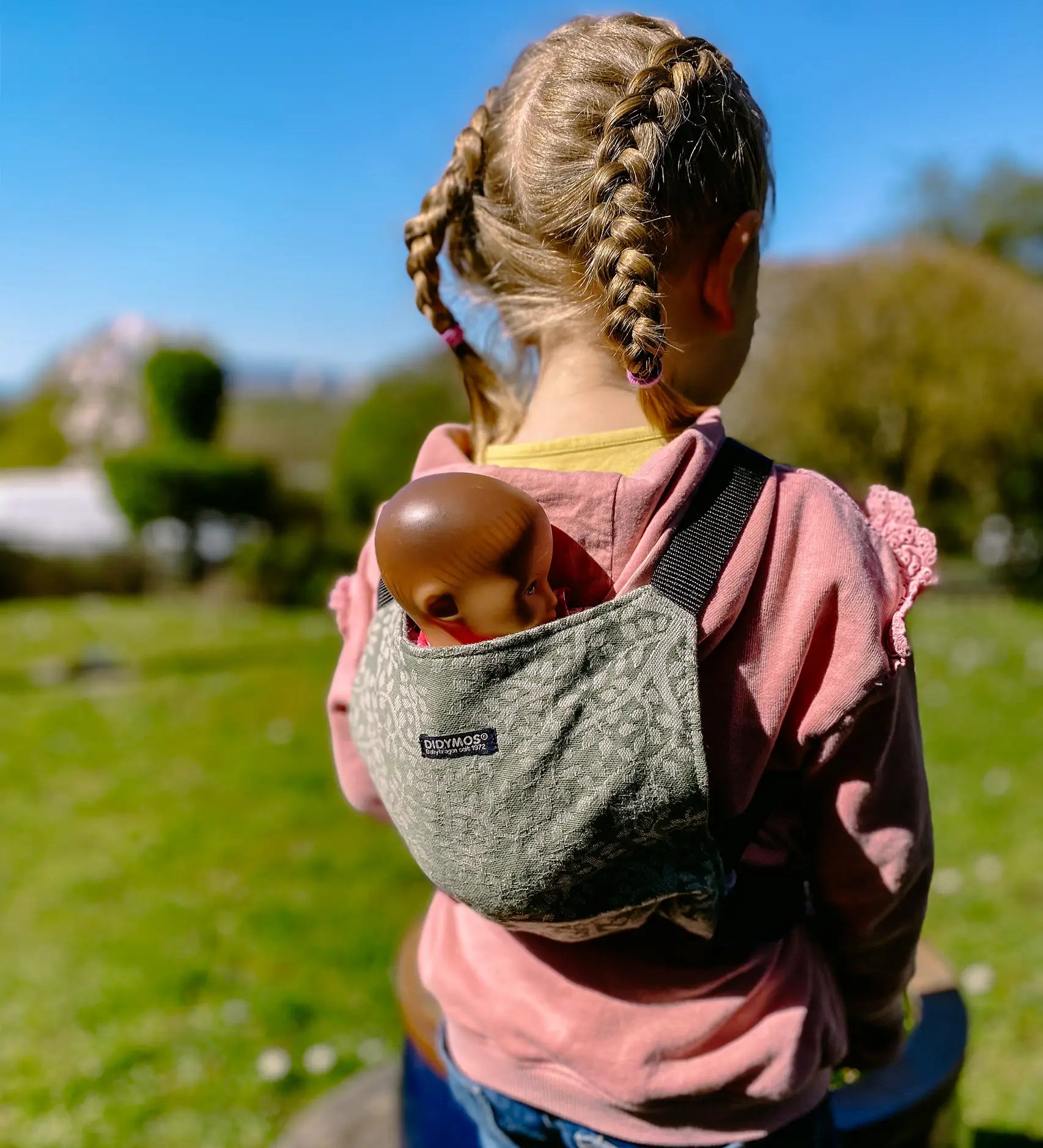 Child carrying a doll in a Didymos toy cotton carrier in olive twig. A green carrier with a repeating tree design. 