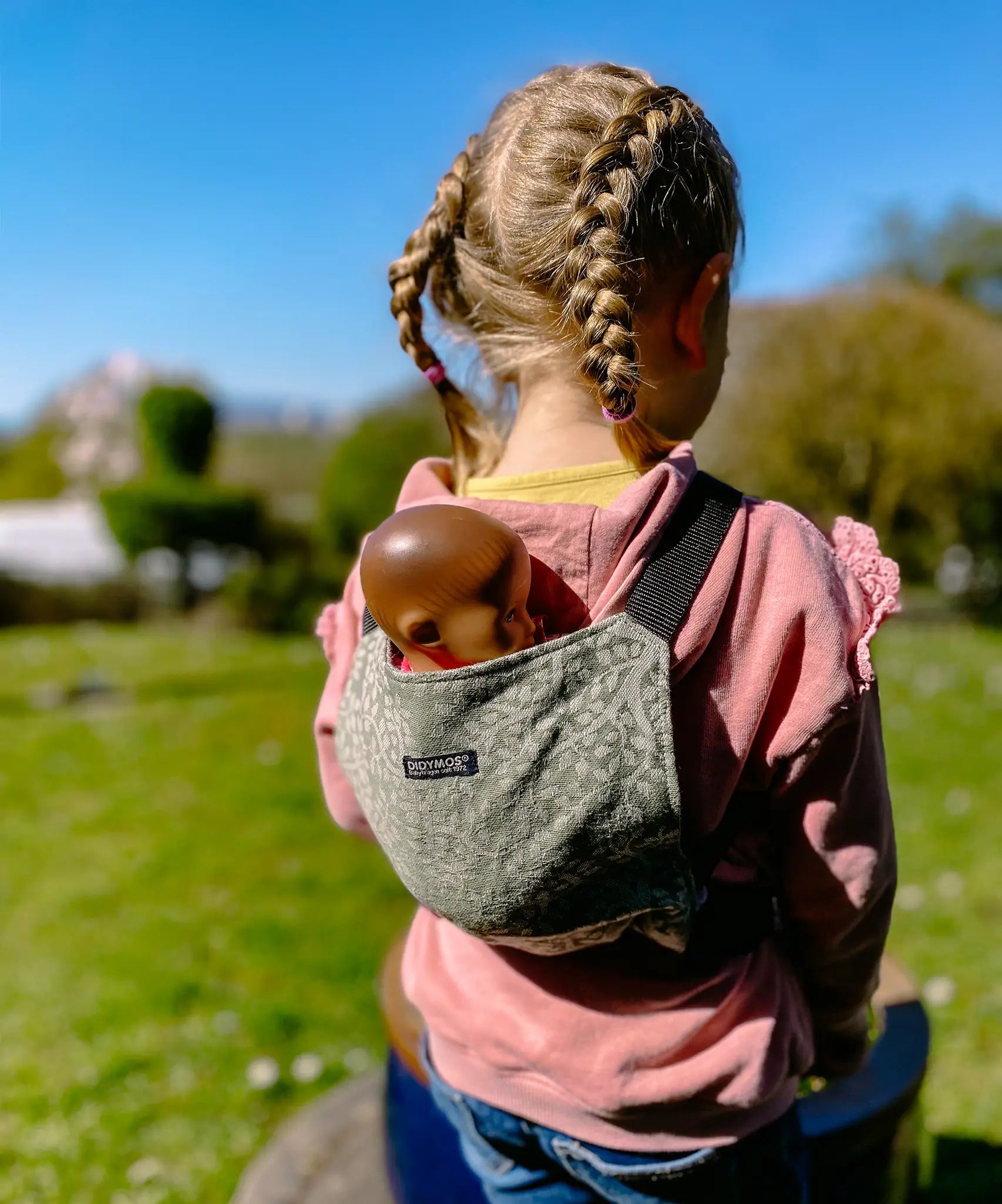 Child carrying a doll in a Didymos toy cotton carrier in olive twig. A green carrier with a repeating tree design. 